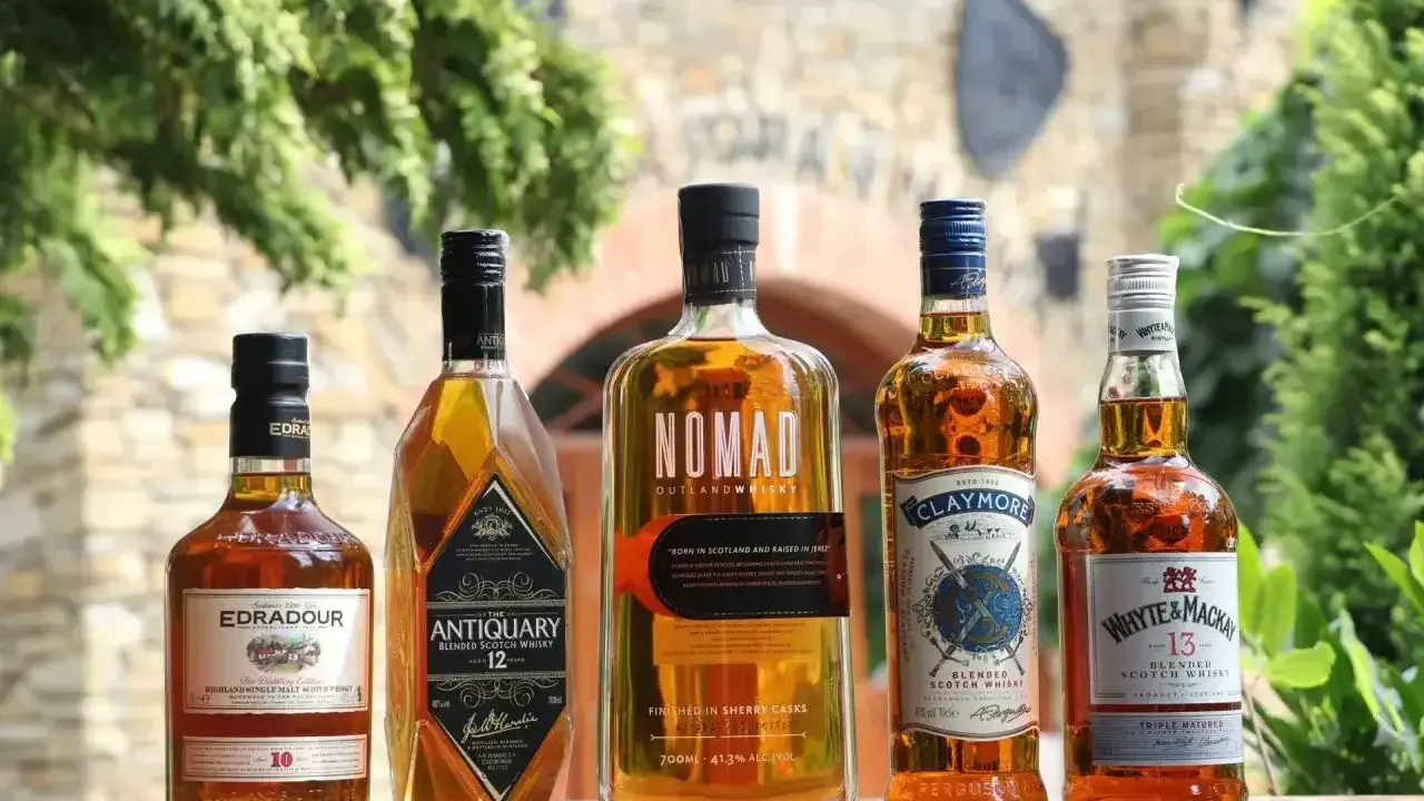 Pięć butelek szkockiej whisky: Edradour, Antiquary, Nomad, Claymore i Whyte & Mackay, stojących na drewnianym stole.