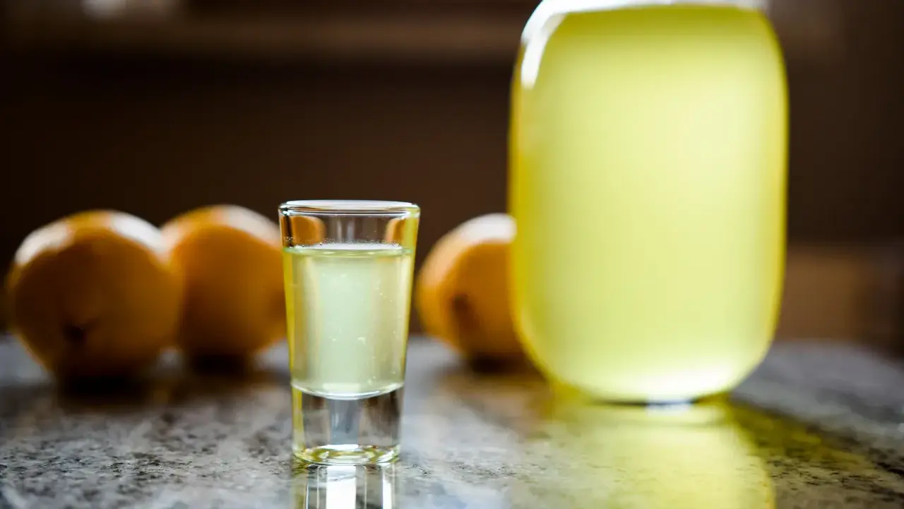 Szklaneczka domowego limoncello, obok cytryny i dużej butelki. Idealny przepis na orzeźwiający trunek.