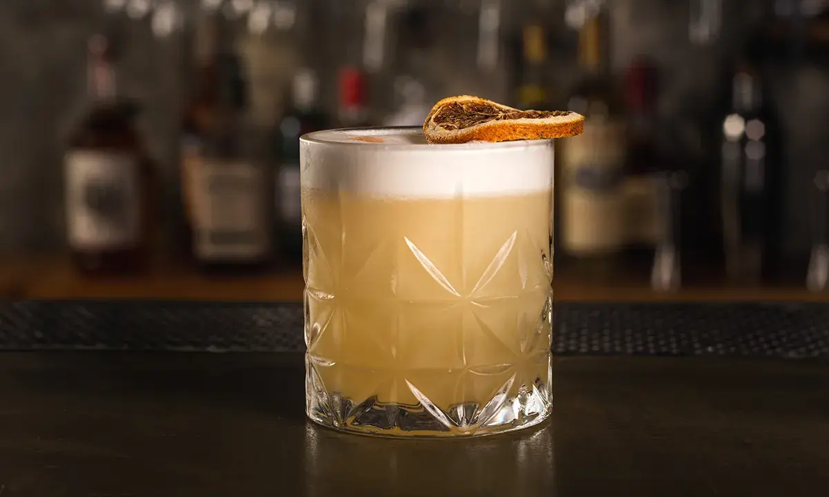 Klasyczny whisky sour z pianką i plasterkiem suszonej pomarańczy, gotowy do degustacji.