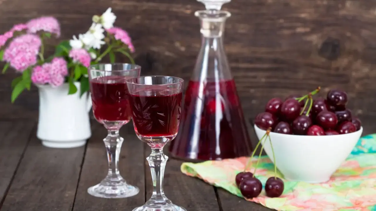 Dwa kieliszki z rubinową nalewką z czereśni na bimbrze, obok miseczka świeżych owoców i bukiecik kwiatów.