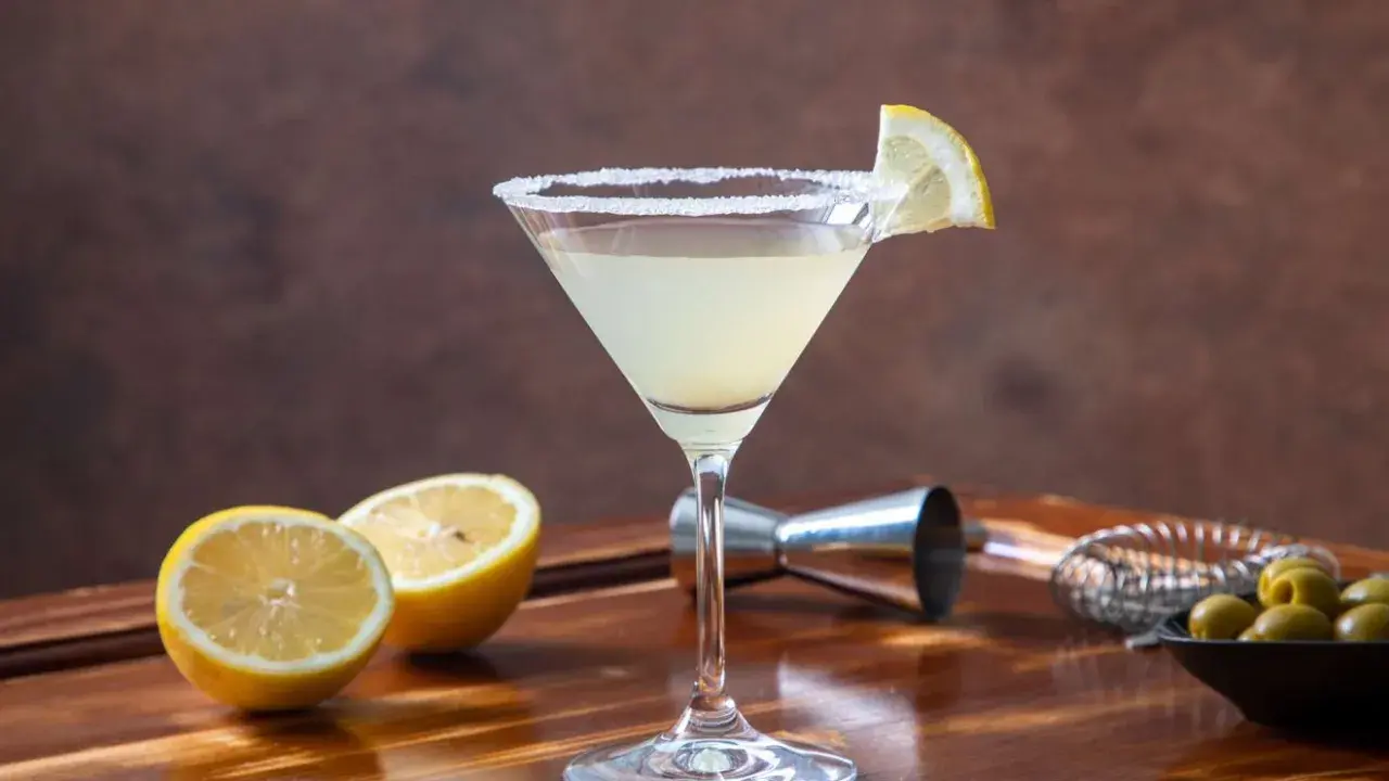 Kieliszek Martini z cytryną i cukrowym brzegiem, obok połówki cytryny, oliwki i akcesoria barmańskie.