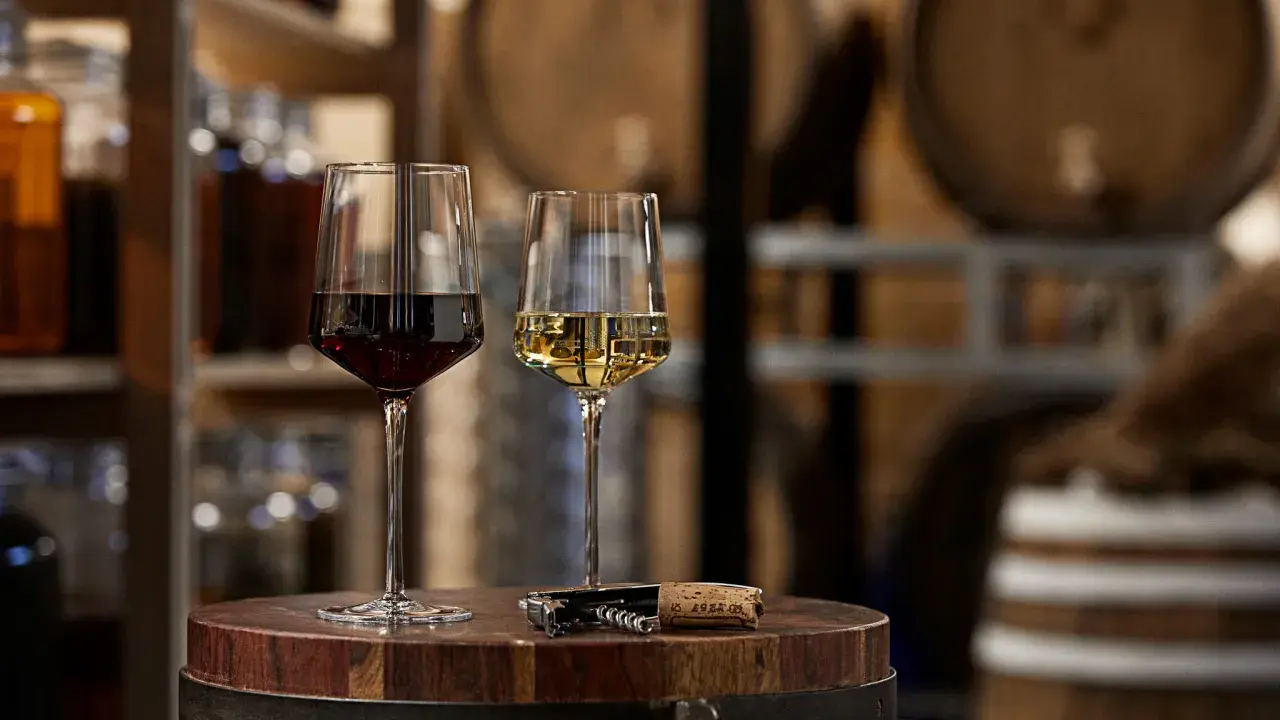 Dwa kieliszki z winem, czerwonym i białym, stoją na drewnianej beczce. Obok leży korkociąg. W tle widać półki z butelkami, być może z grappa alkohol.