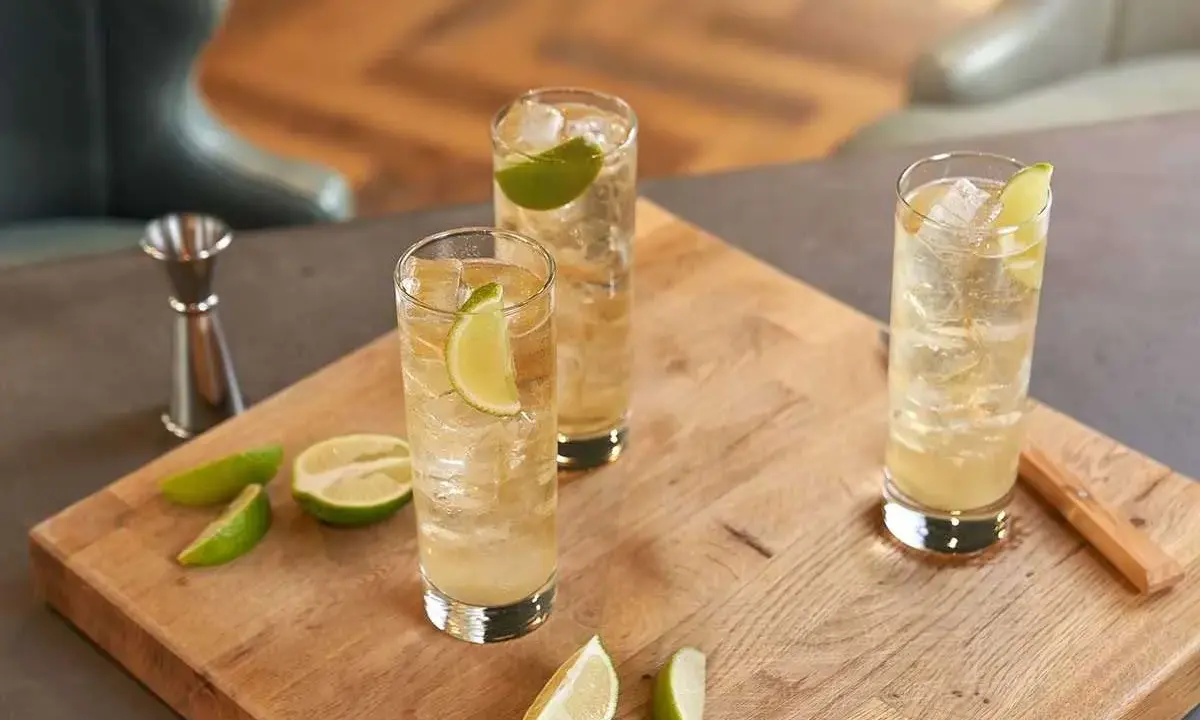 Trzy orzeźwiające whisky ze spritem, udekorowane plasterkami limonki, stoją na drewnianej desce. Obok leżą świeże kawałki limonki.