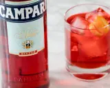 Butelka Campari i szklanka z czerwonym drinkiem, lodem i skórką pomarańczy. Idealny aperitif, który może być bazą dla drinków z Aperolem.