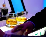 Dwa kieliszki złocistej whisky na barze, idealne na dzień whisky. Dłoń w garniturze sięga po drinka.