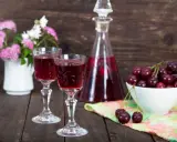 Dwa kieliszki z rubinową nalewką z czereśni na bimbrze, obok miseczka świeżych owoców i bukiecik kwiatów.