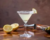 Kieliszek Martini z cytryną i cukrowym brzegiem, obok połówki cytryny, oliwki i akcesoria barmańskie.