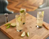 Trzy orzeźwiające whisky ze spritem, udekorowane plasterkami limonki, stoją na drewnianej desce. Obok leżą świeże kawałki limonki.