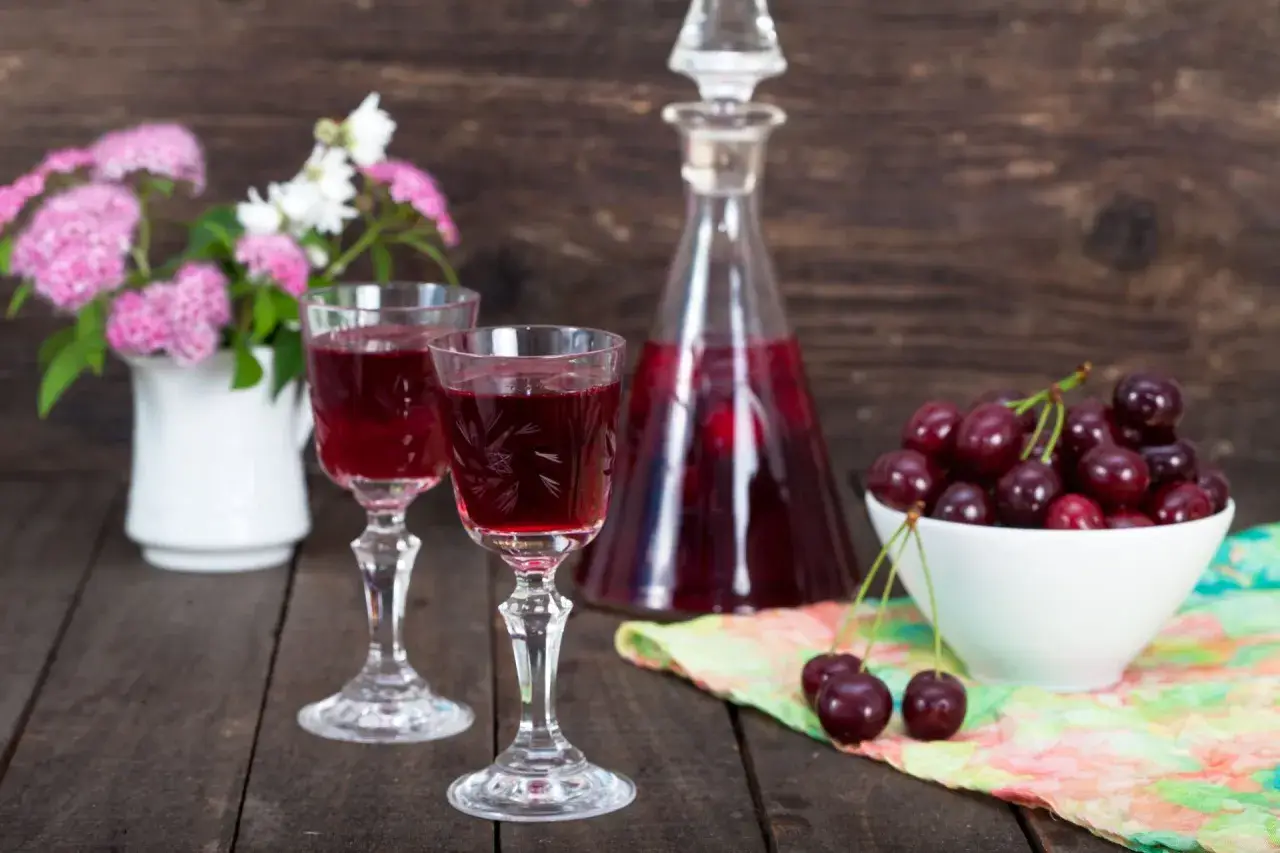 Dwa kieliszki z rubinową nalewką z czereśni na bimbrze, obok miseczka świeżych owoców i bukiecik kwiatów.
