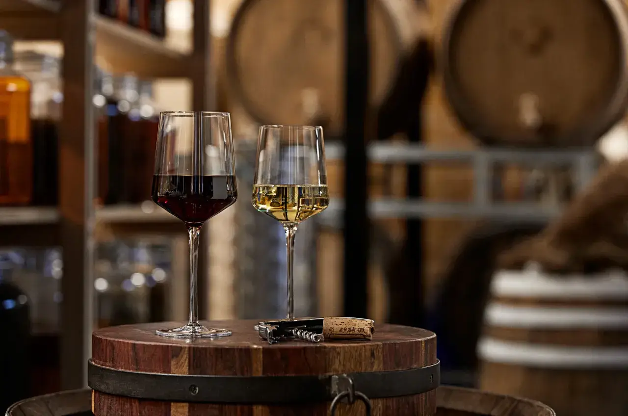Dwa kieliszki z winem, czerwonym i białym, stoją na drewnianej beczce. Obok leży korkociąg. W tle widać półki z butelkami, być może z grappa alkohol.