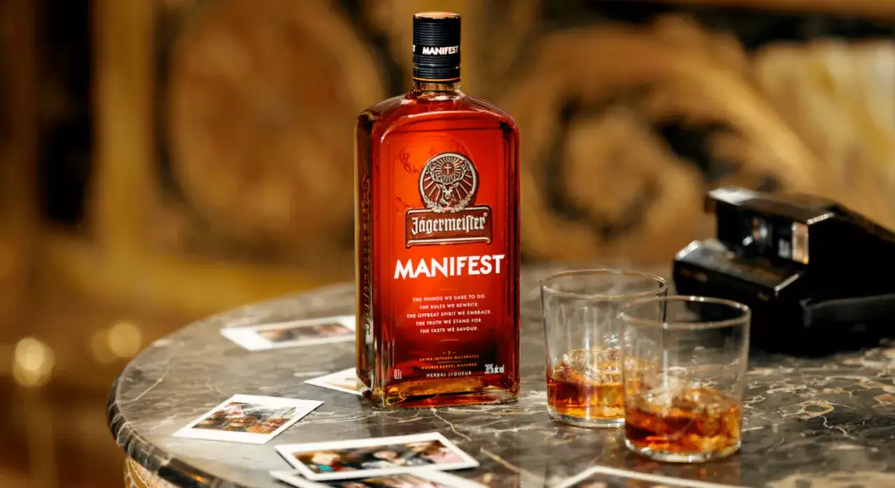 Jägermeister Manifest z czym pić? Butelka i dwa kieliszki z bursztynowym trunkiem na marmurowym blacie, obok starego aparatu i zdjęć.