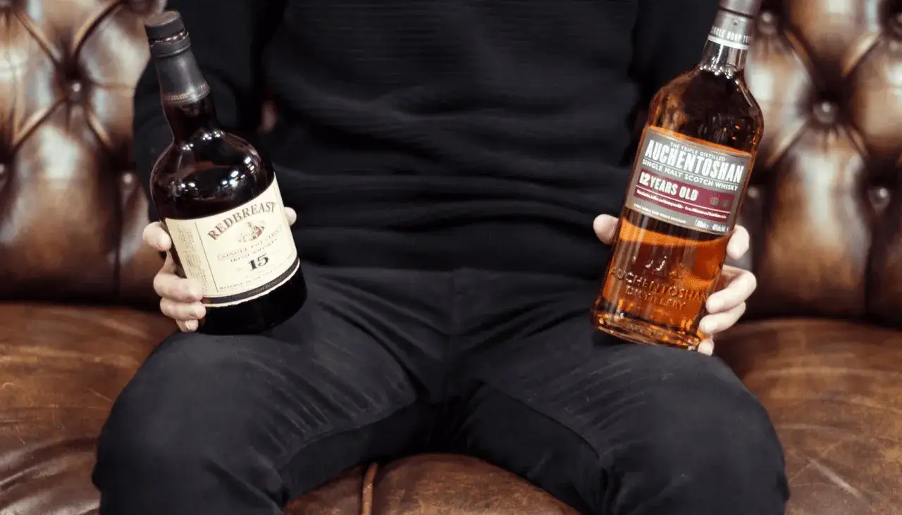 Osoba trzyma dwie butelki: Redbreast 15 Year Old (irlandzka whisky) i Auchentoshan 12 Year Old (szkocka whisky).