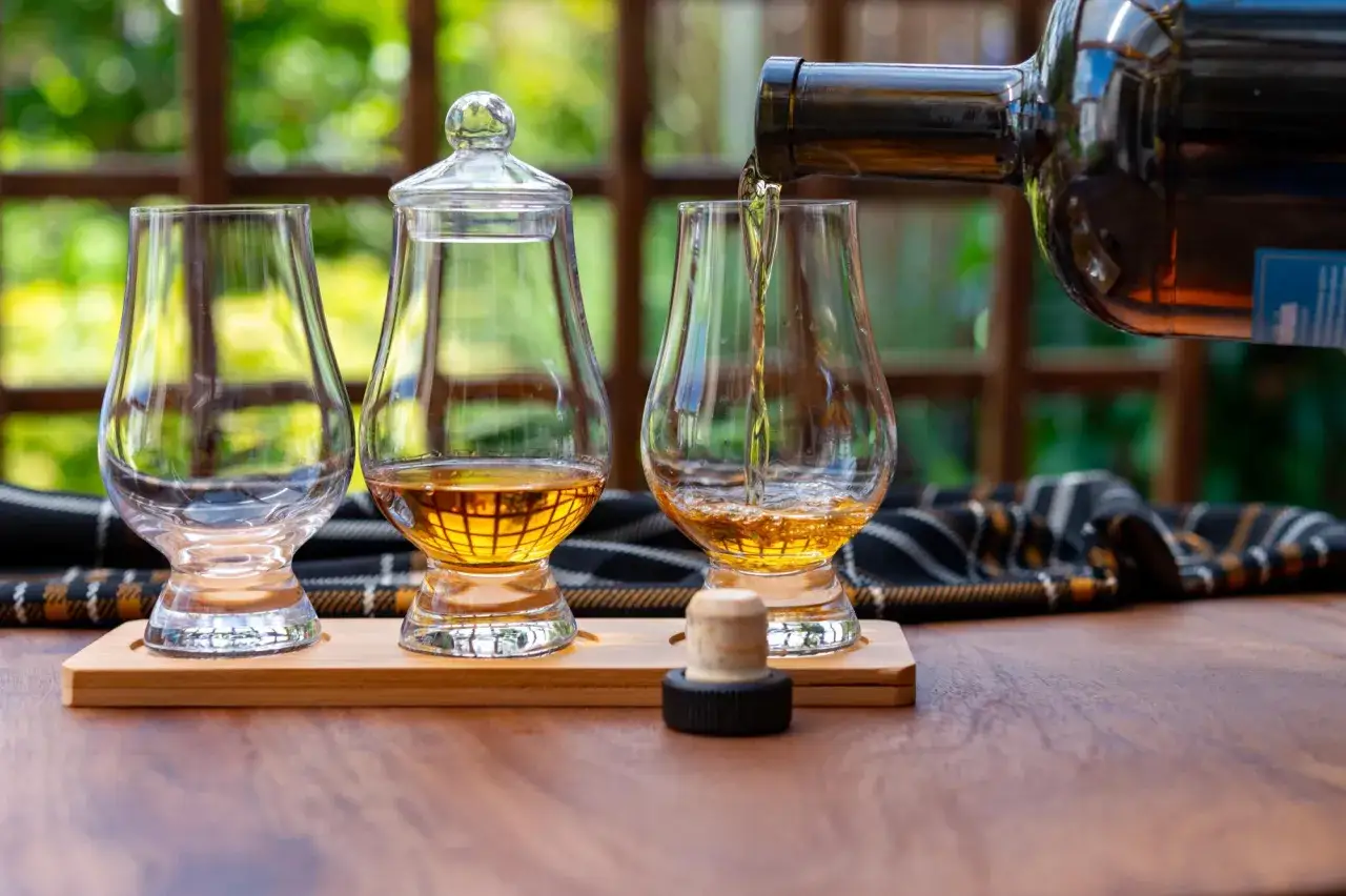 Degustacja burbonu i whisky. Bursztynowy trunek nalewany z butelki do kieliszk&oacute;w na drewnianej podstawce.