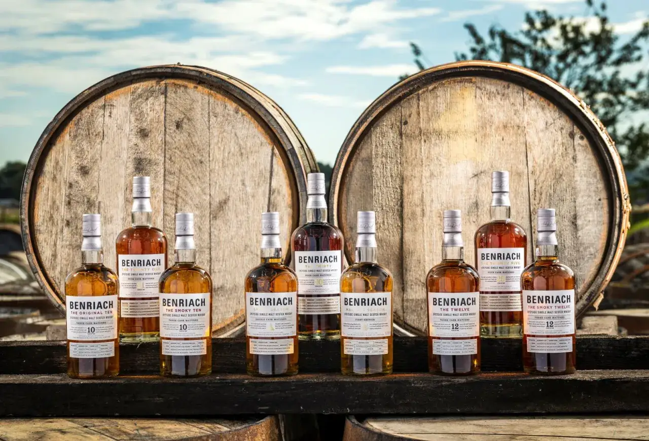Butelki whisky BenRiach stoją przed beczkami, pokazując, skąd pochodzi whisky.