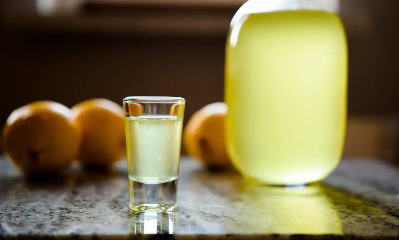 Szklaneczka orzeźwiającego limoncello obok cytryn i dużej butelki. Idealny przepis na lato.