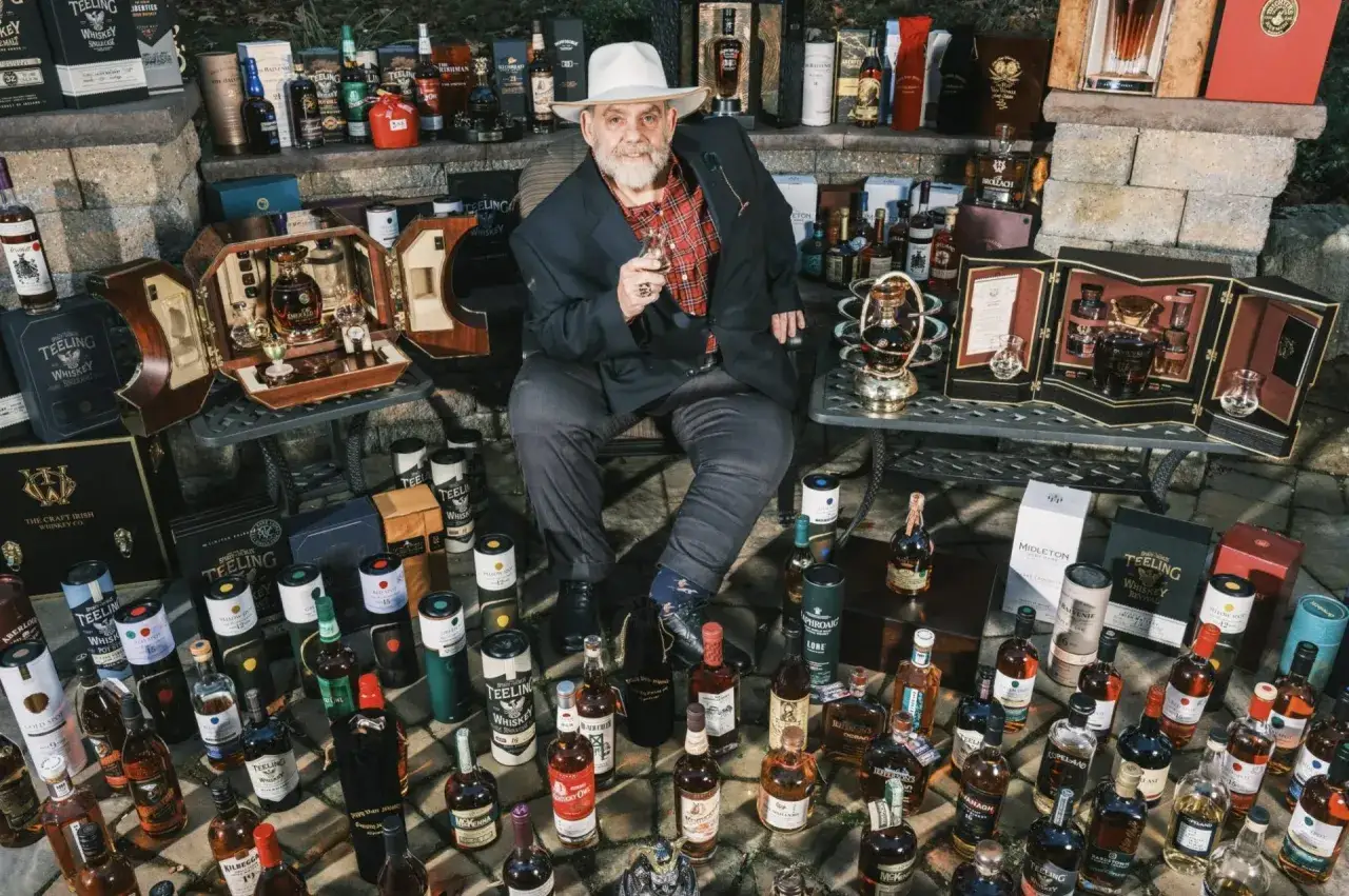 Mężczyzna w kapeluszu siedzi wśród kolekcji drogich whisky, trzymając kieliszek.